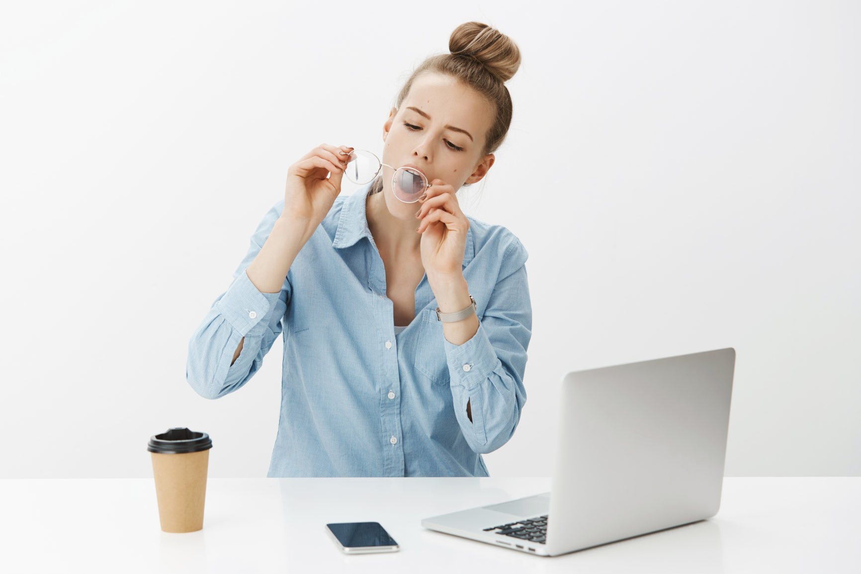 Girl breathing on glasses as cleaning lens from dust, whiping eyewear, being lazy not doing work as sitting in office near laptop and smartphone, drinking coffee, skipping job over gray background.